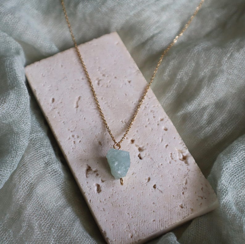 May include: A delicate gold chain necklace featuring a pale blue aquamarine gemstone pendant. The raw, irregular shape of the stone contrasts with the smooth, polished chain. The necklace rests on a textured, rectangular stone against a soft green backdrop.