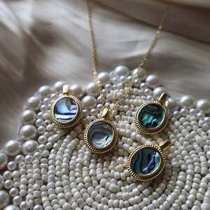 May include: A gold-toned necklace and three matching pendants, each featuring a circular abalone shell inlay with iridescent blue and green hues, framed by a textured beaded border and a small bail at the top for attachment to a chain.