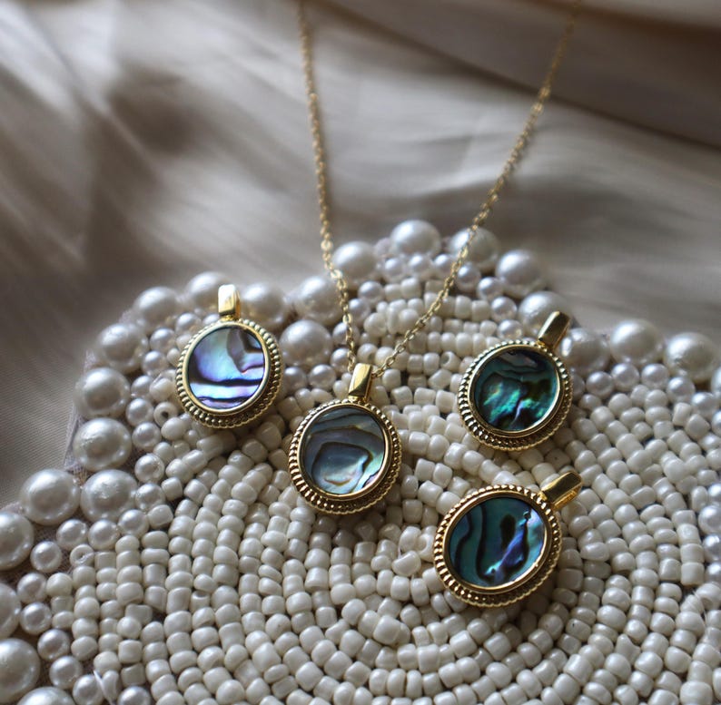 May include: A gold-toned necklace and three matching pendants, each featuring a circular abalone shell inlay with iridescent blue and green hues, framed by a textured beaded border and a small bail at the top for attachment to a chain.