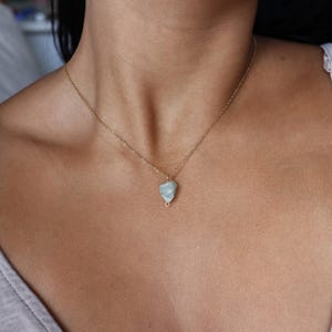 May include: A delicate gold-colored chain necklace featuring a small, light blue gemstone pendant. The pendant has a rough, natural texture and is wire-wrapped. The necklace is worn around the neck.