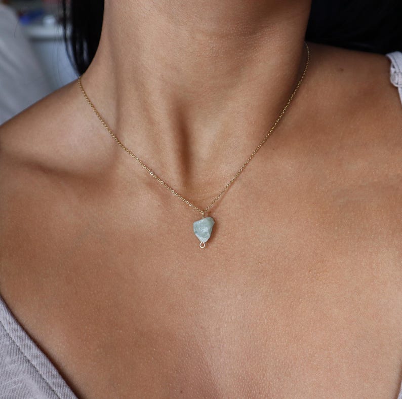 May include: A delicate gold-colored chain necklace featuring a small, light blue gemstone pendant. The pendant has a rough, natural texture and is wire-wrapped. The necklace is worn around the neck.