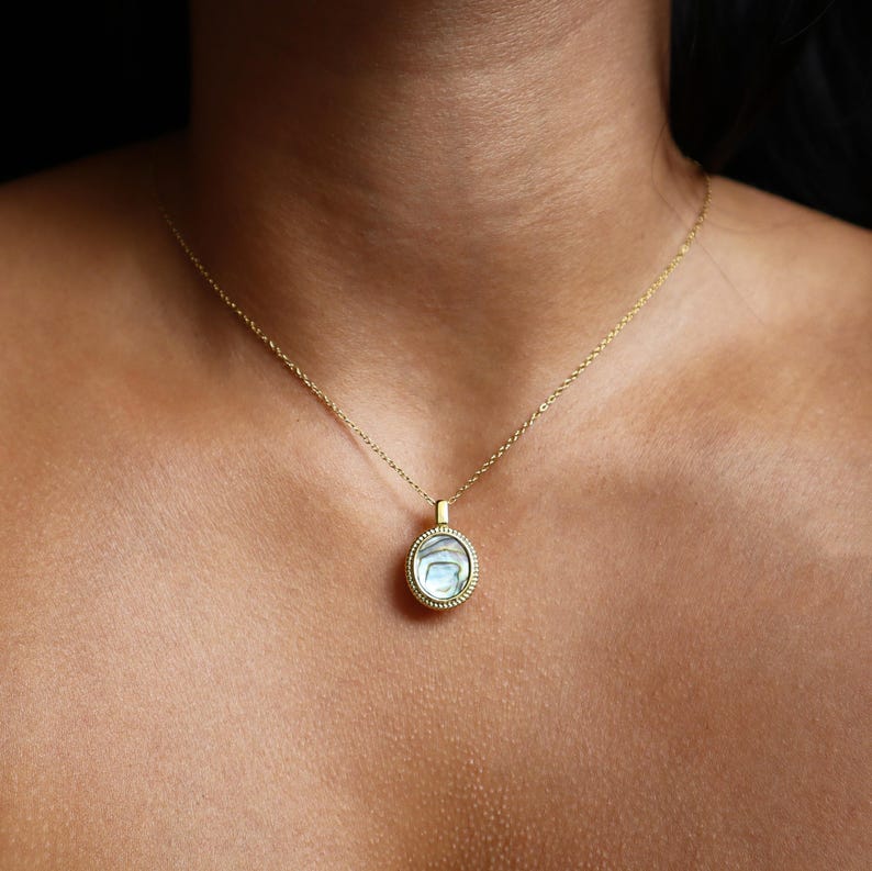May include: A gold-toned necklace featuring a circular pendant with an iridescent abalone shell inlay. The pendant is framed by a textured, beaded metal border and hangs from a delicate, thin gold-toned chain that rests against the collarbone.