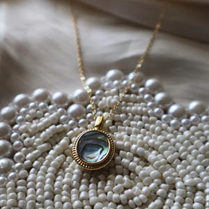 May include: A gold-toned necklace featuring a circular pendant with a textured, beaded edge and a central inlay of iridescent abalone shell. The pendant is suspended from a delicate gold-toned chain, showcasing a polished finish and reflective surface.