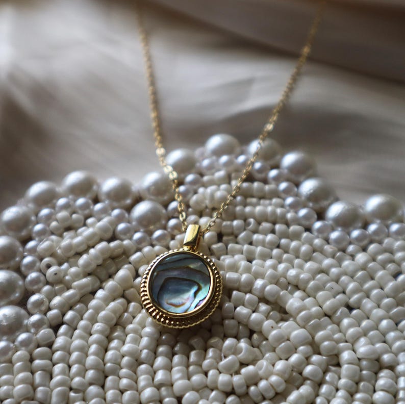 May include: A gold-toned necklace featuring a circular pendant with a textured, beaded edge and a central inlay of iridescent abalone shell. The pendant is suspended from a delicate gold-toned chain, showcasing a polished finish and reflective surface.