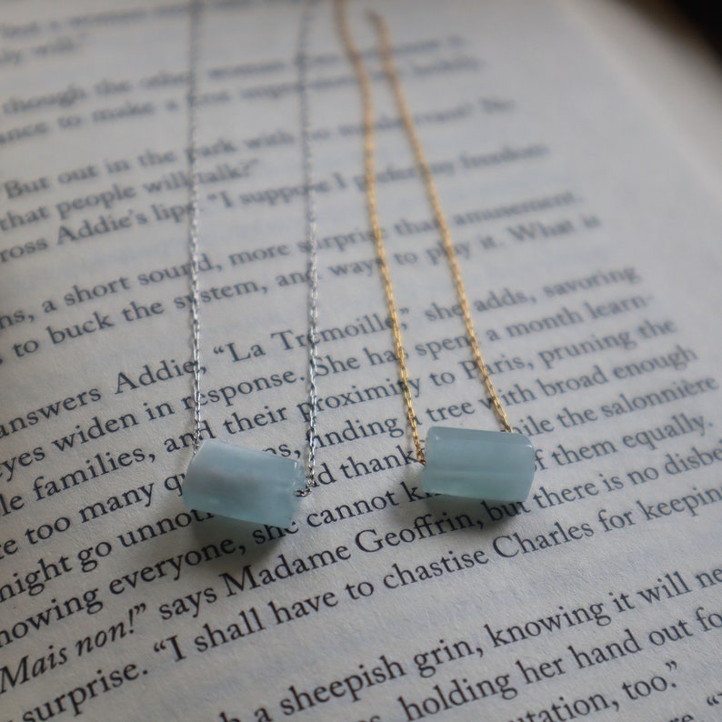 May include: Three delicate necklaces with light blue gemstone beads. The necklaces are layered on a book with text visible in the background.