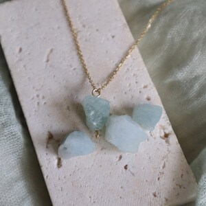 May include: A gold-colored chain necklace featuring a cluster of light blue, raw aquamarine stones. The necklace is displayed on a textured, off-white surface, with a soft green fabric in the background. The aquamarine stones have a natural, irregular shape.