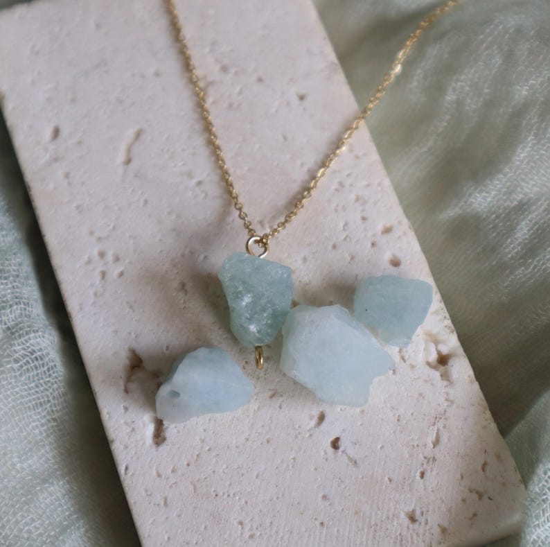May include: A gold-colored chain necklace featuring a cluster of light blue, raw aquamarine stones. The necklace is displayed on a textured, off-white surface, with a soft green fabric in the background. The aquamarine stones have a natural, irregular shape.