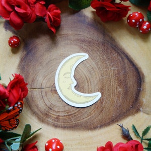 May include: A yellow crescent moon sticker with a closed eye and a smiling mouth. The sticker is on a wooden surface with red flowers and mushrooms.