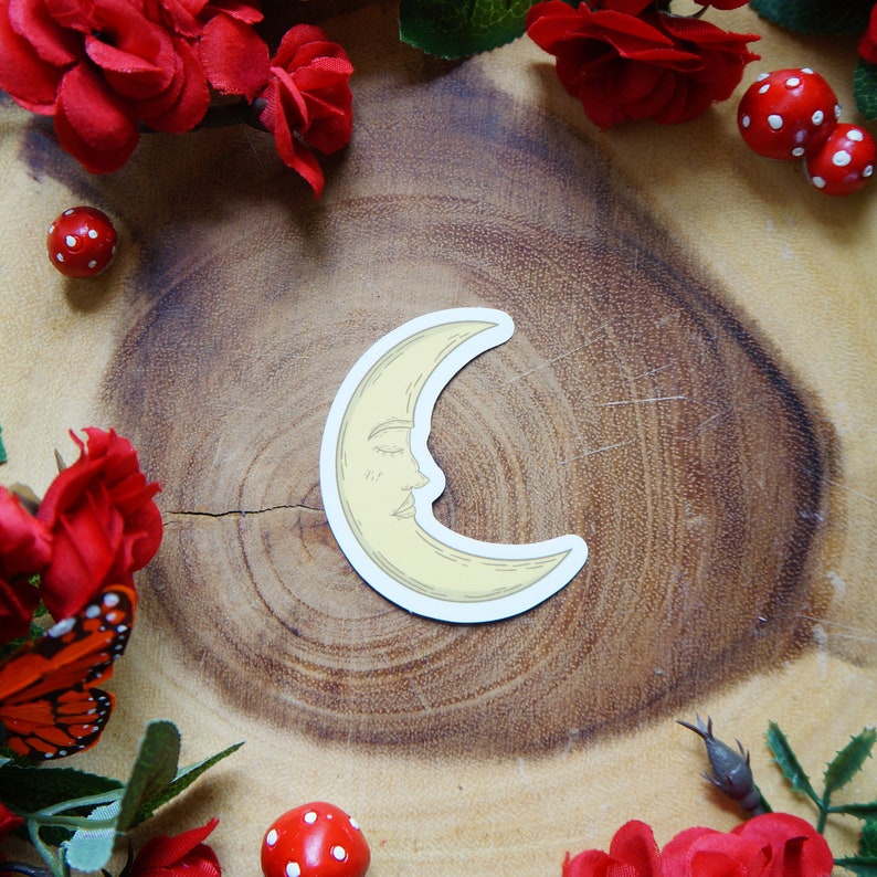 May include: A yellow crescent moon sticker with a closed eye and a smiling mouth. The sticker is on a wooden surface with red flowers and mushrooms.