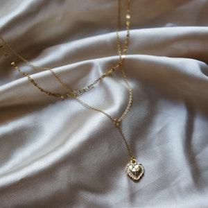 May include: A gold layered necklace with a heart pendant. The pendant has a textured surface and is adorned with small sparkling stones.