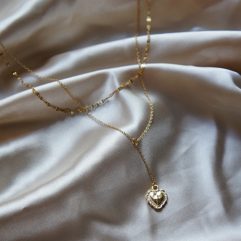 May include: A gold layered necklace with a heart pendant. The pendant has a textured surface and is adorned with small sparkling stones.