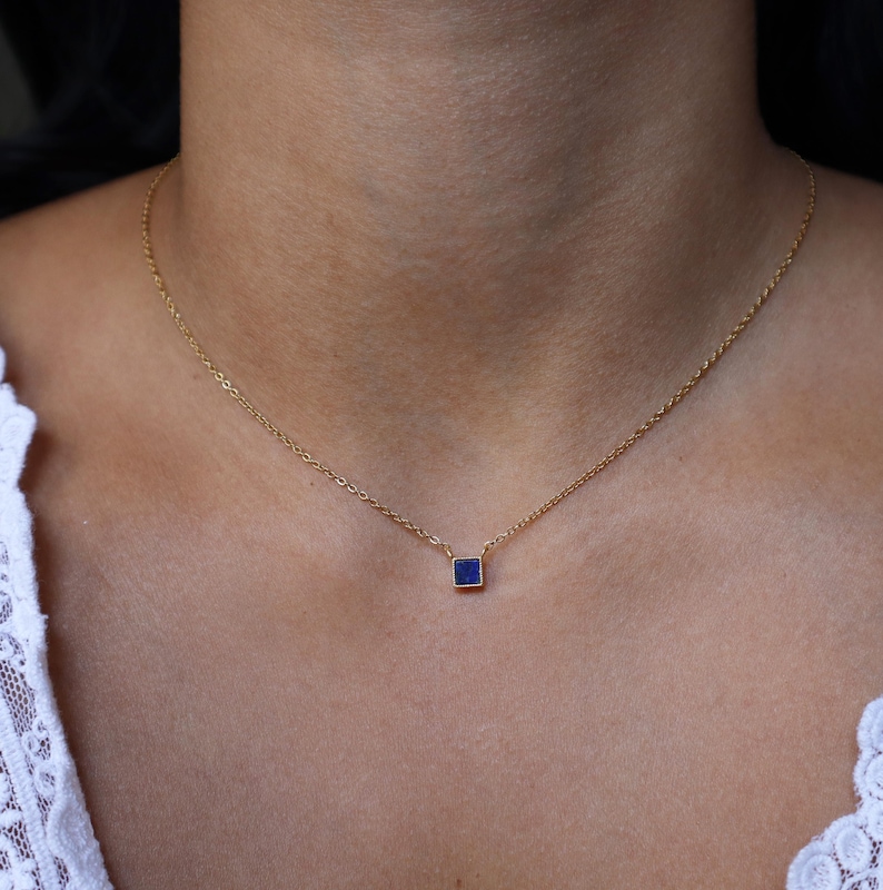Peut inclure: Un collier d&eacute;licat en cha&icirc;ne dor&eacute;e avec un petit pendentif de forme carr&eacute;e orn&eacute; d'une pierre pr&eacute;cieuse bleu fonc&eacute;.