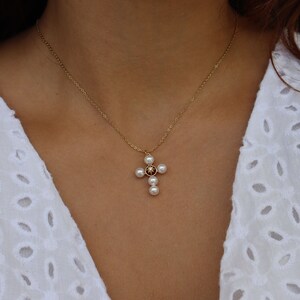 Puede incluir: Un collar de cadena dorada con un colgante de cruz de perlas. La cruz tiene una peque&ntilde;a piedra preciosa roja oscura en el centro.