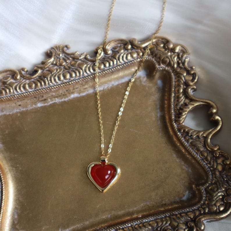 May include: A gold chain necklace with a red heart pendant. The pendant has a gold frame and a red stone in the center.
