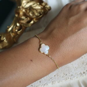 May include: A gold-colored chain bracelet featuring a four-leaf clover charm with a white, pearlescent center. The bracelet is delicate and appears to be worn on a wrist. The background includes a gold-colored decorative object.