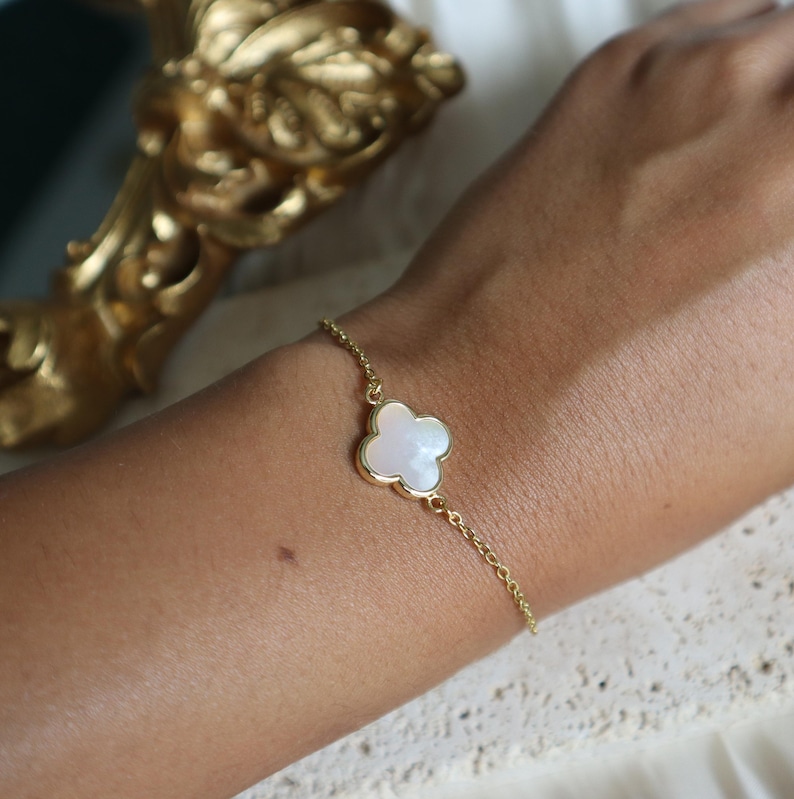 May include: A gold-colored chain bracelet featuring a four-leaf clover charm with a white, pearlescent center. The bracelet is delicate and appears to be worn on a wrist. The background includes a gold-colored decorative object.