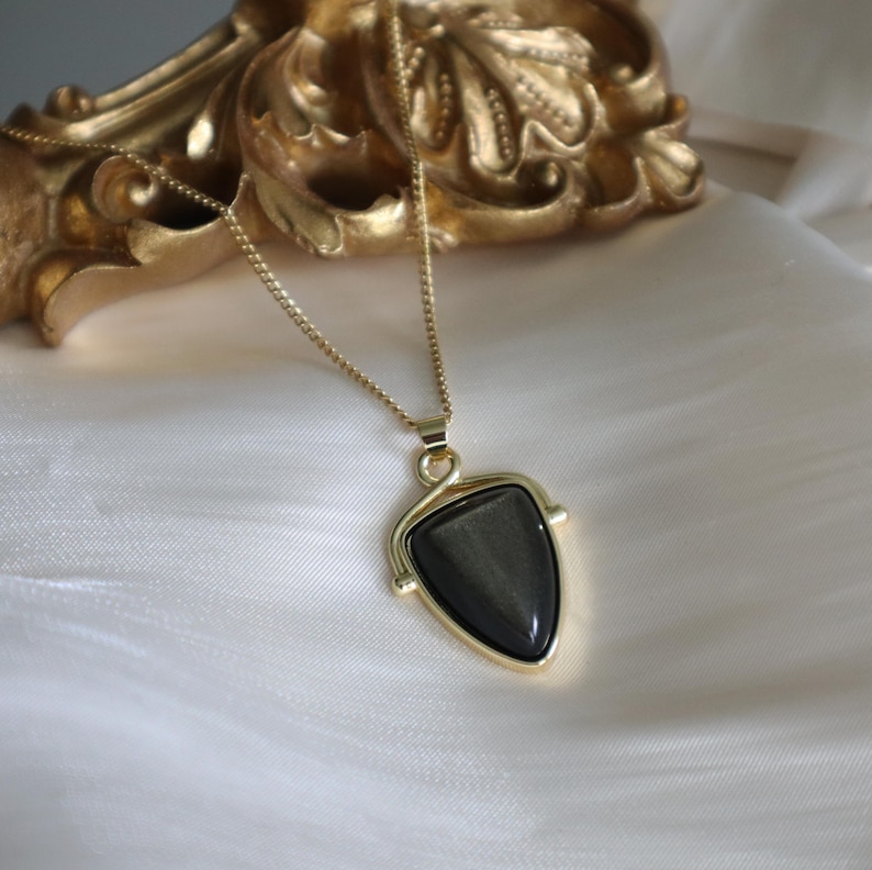 May include: A gold chain necklace with a black gemstone pendant. The pendant is shaped like a triangle with a gold frame.