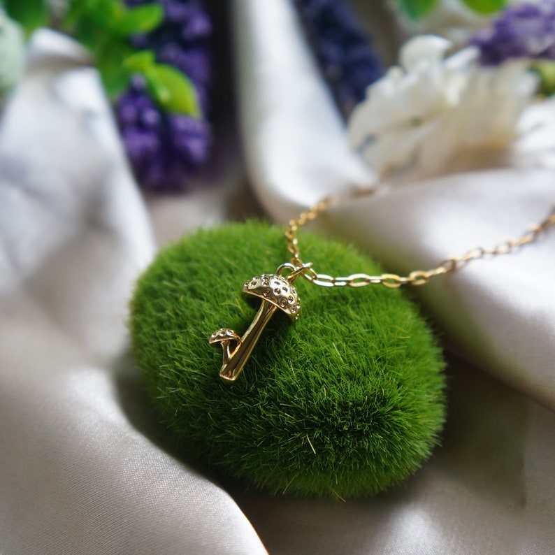 Puede incluir: Un collar con colgante de seta dorado. El colgante presenta peque&ntilde;os detalles decorativos en el sombrero y el tallo. El collar se muestra sobre musgo verde, con un fondo satinado.