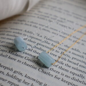 May include: Two delicate gold chains with light blue gemstone beads. The chains are layered on top of a book with text.