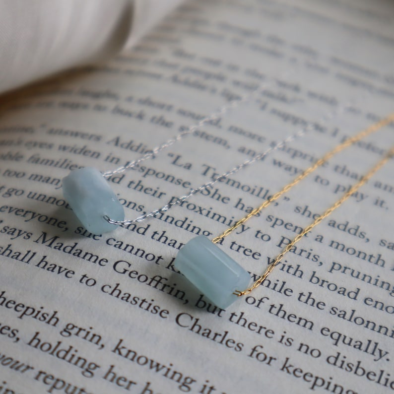 May include: Two delicate gold chains with light blue gemstone beads. The chains are layered on top of a book with text.