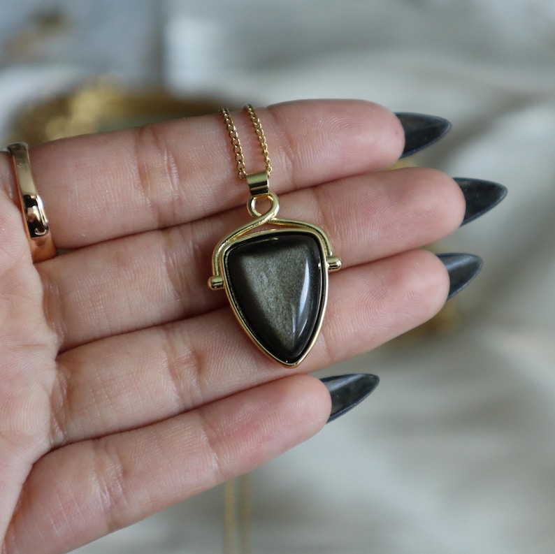 May include: A gold chain necklace with a spinning triangular pendant featuring a dark gray stone with a shimmery effect.