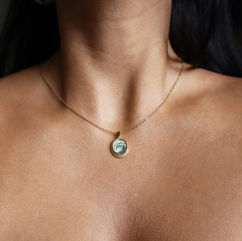 May include: A gold-toned necklace featuring a circular pendant with a textured, rope-style border. The centre of the pendant contains an iridescent, swirling abalone shell inlay with shades of blue, green, and grey, suspended on a thin gold chain.