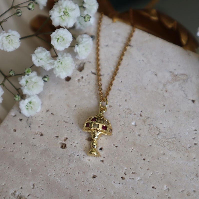 May include: A gold necklace with a pendant shaped like a lamp with red jewels. The lamp is detailed with a geometric design.