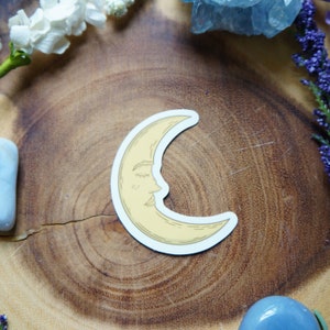 May include: A yellow crescent moon sticker with a closed eye and a nose, on a wooden surface.