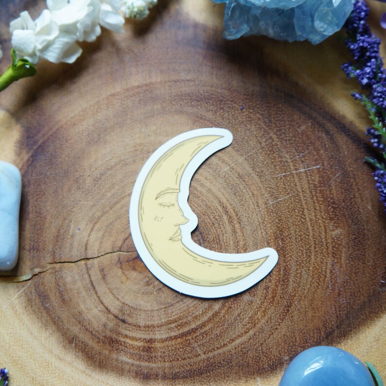 May include: A yellow crescent moon sticker with a closed eye and a nose, on a wooden surface.