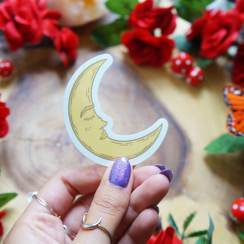 May include: A yellow crescent moon sticker with a closed eye and a nose, on a white background. The sticker is being held by a hand with purple nail polish.