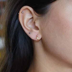 May include: Gold hoop earrings with a small, white opal evil eye charm.