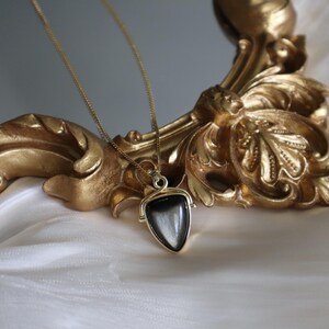 May include: A gold chain necklace with a black gemstone pendant. The pendant is shaped like a triangle and has a gold frame. The necklace is resting on a white surface.