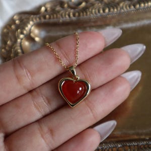 May include: A gold chain necklace with a heart-shaped pendant. The pendant has a red carnelian stone set in a gold frame.