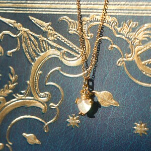 May include: A gold necklace with a delicate pendant shaped like a heart. The pendant is made of white opal and has a gold filigree design. The necklace is photographed on a blue and gold patterned surface.