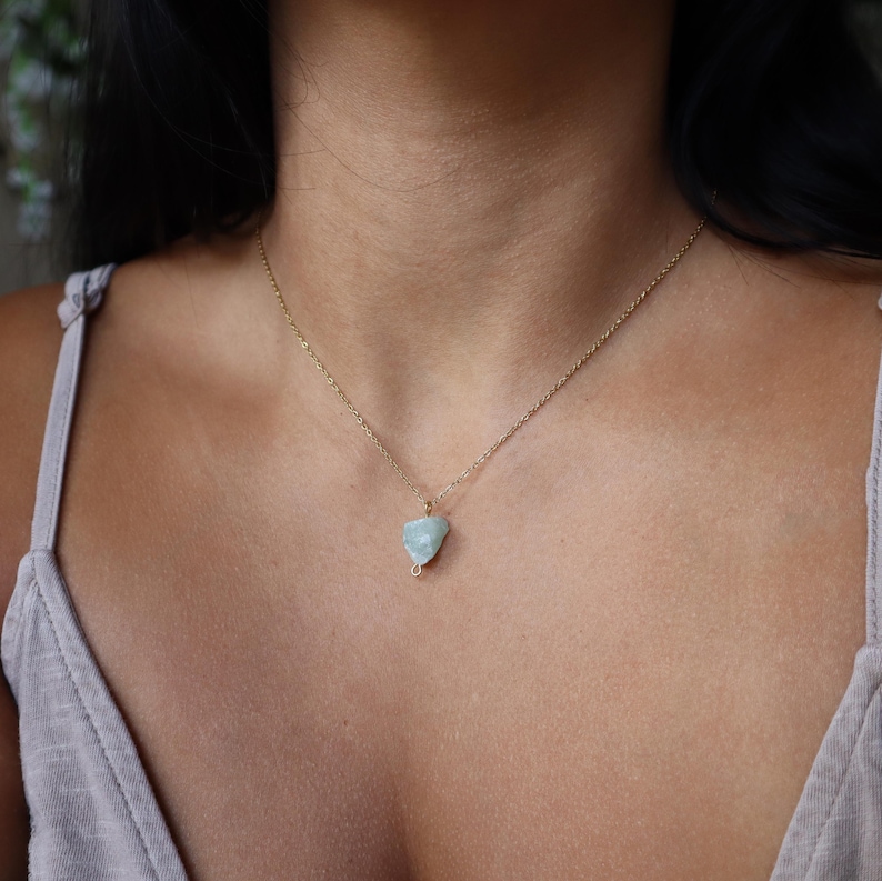May include: A delicate gold chain necklace featuring a pale blue-green gemstone pendant. The pendant is a rough, irregular shape, and the necklace is displayed against a neutral background.