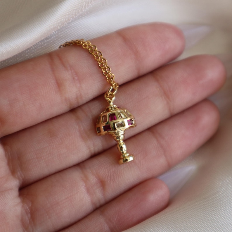 May include: A gold-tone necklace with a lamp-shaped pendant. The pendant is decorated with clear and red cubic zirconia stones.