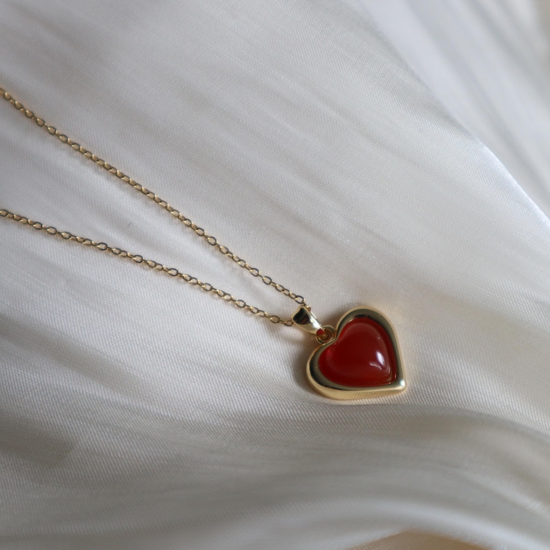 May include: A gold chain necklace with a red heart pendant. The heart is made of a red gemstone and has a gold border.