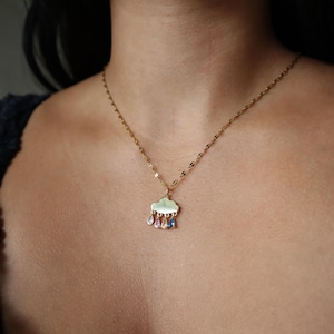May include: A gold necklace with a cloud pendant and three colourful teardrop gems. The pendant features pink, purple, and blue gems. The necklace is worn around the neck and has a delicate chain.