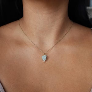 May include: A delicate gold-tone necklace featuring a small, pale blue-green gemstone pendant. The pendant is irregularly shaped and wire-wrapped, suspended from a fine chain. The necklace is shown against a skin tone background.