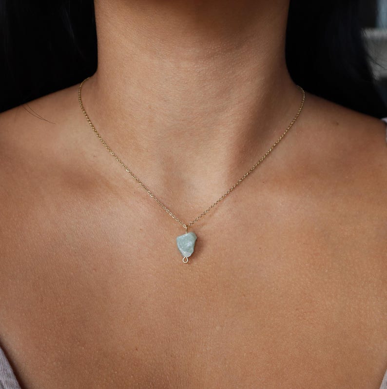 May include: A delicate gold-tone necklace featuring a small, pale blue-green gemstone pendant. The pendant is irregularly shaped and wire-wrapped, suspended from a fine chain. The necklace is shown against a skin tone background.