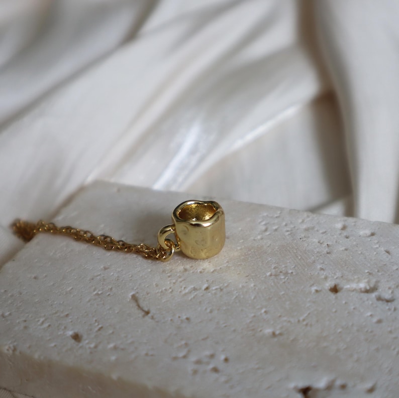May include: A gold-toned necklace with a small, textured mug charm. The mug charm is a simple design with a rounded handle.