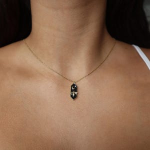 May include: A gold chain necklace with a black enamel pendant featuring a crescent moon and a bee. The pendant is shaped like a long, thin rectangle with rounded edges.