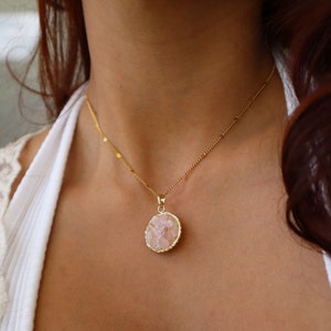 May include: A gold chain necklace with a pink crystal pendant. The pendant is a round, flat piece of crystal with a gold rim.