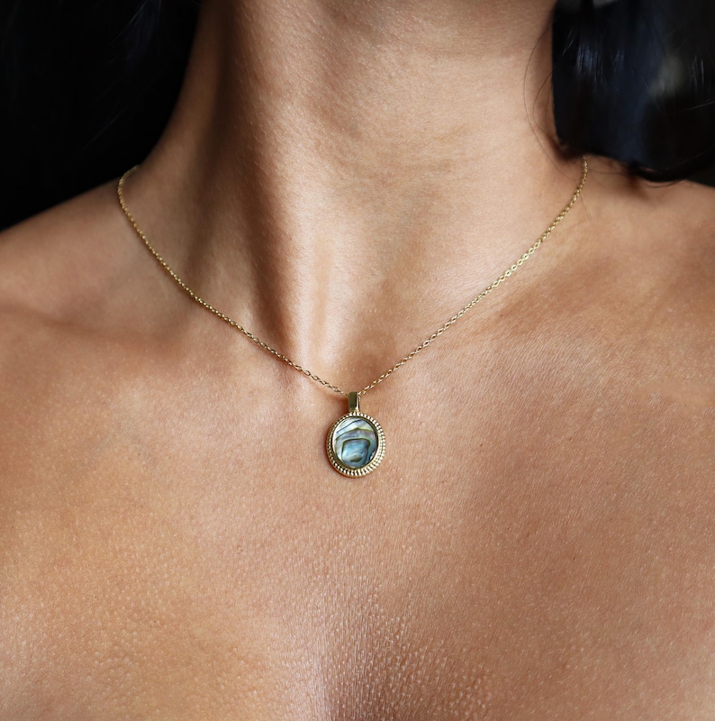 May include: A gold-toned necklace featuring a circular pendant with a textured, beaded edge. The centre of the pendant contains an iridescent, swirling abalone shell inlay with blue, green, and grey tones, suspended from a delicate gold-toned chain.