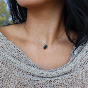 May include: A delicate gold chain necklace featuring a dark green, oval-shaped gemstone pendant. The necklace is worn against a neutral-toned top, creating a simple and elegant look. The gemstone has a subtle, natural sheen.