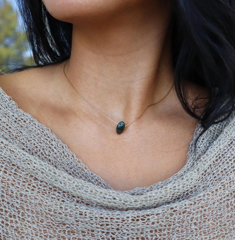 May include: A delicate gold chain necklace featuring a dark green, oval-shaped gemstone pendant. The necklace is worn against a neutral-toned top, creating a simple and elegant look. The gemstone has a subtle, natural sheen.