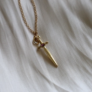 May include: A gold-colored pendant necklace featuring a small sword design with a pink gemstone in the center. The sword pendant hangs from a delicate gold chain, set against a soft, off-white background.