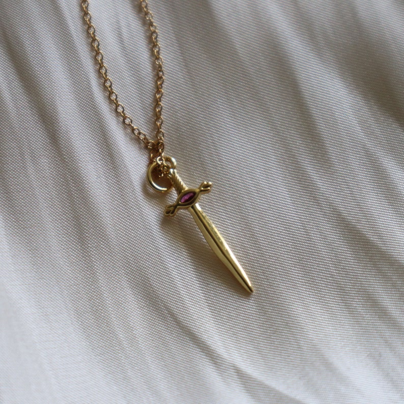 May include: A gold-colored pendant necklace featuring a small sword design with a pink gemstone in the center. The sword pendant hangs from a delicate gold chain, set against a soft, off-white background.
