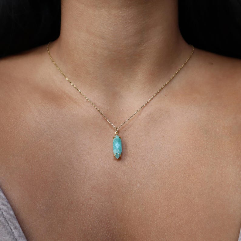 K&ouml;nnte beinhalten: Eine zarte goldene Halskette mit einem Anh&auml;nger aus hellblauem, facettiertem Edelstein. Der Anh&auml;nger hat eine l&auml;ngliche ovale Form. Die Halskette wird um den Hals getragen.