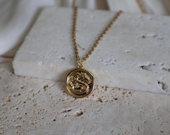 May include: A gold-colored necklace featuring a circular pendant with a detailed dragon design. The pendant hangs from a delicate chain. The necklace is displayed on a textured, off-white surface, creating a contrast with the shiny gold.